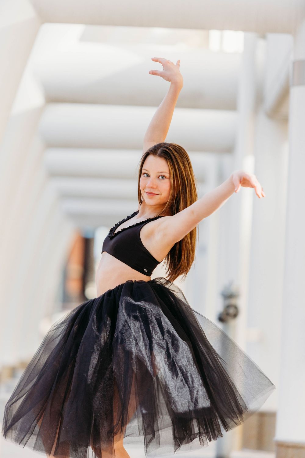 Dancer in black tutu skirt and crop top performs graceful ballet pose in bright white studio setting.