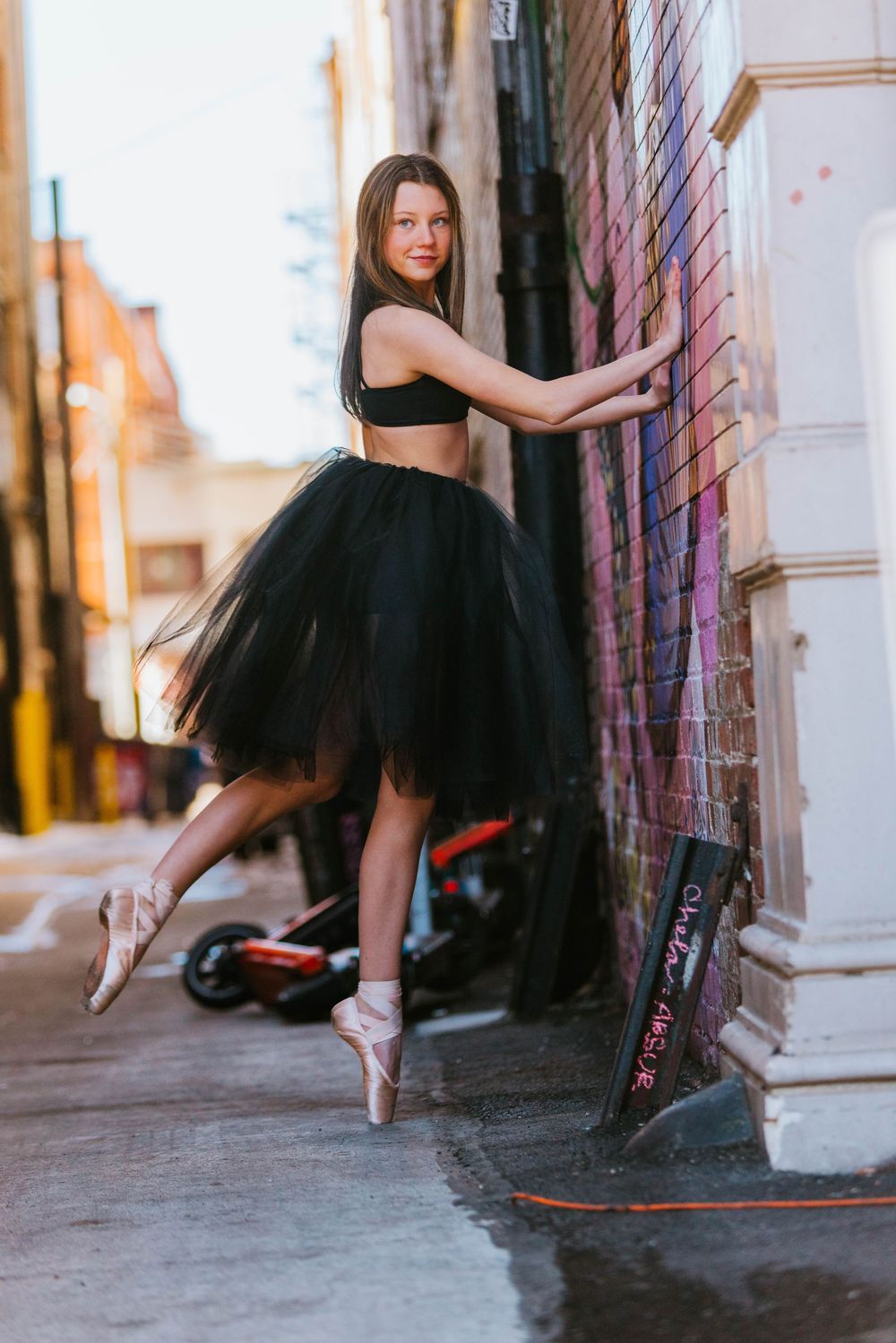 Ballet dancer in black tulle tutu dress poses gracefully in urban alleyway with graffiti walls.