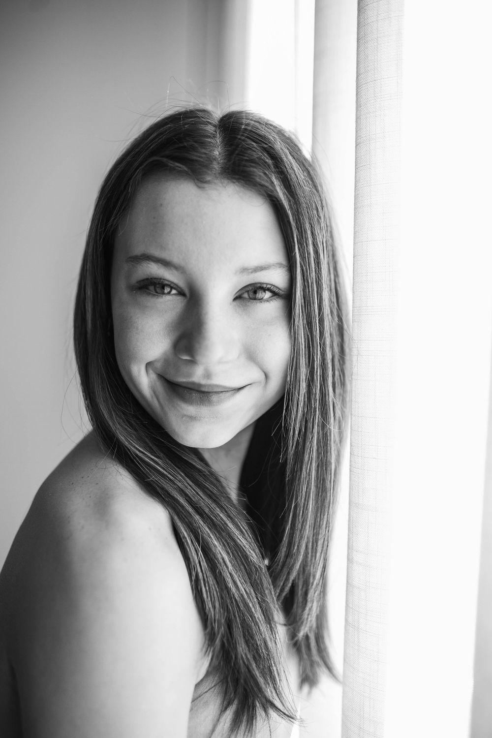 Black and white portrait photograph showing a person with long straight dark hair and a gentle smile near a window.