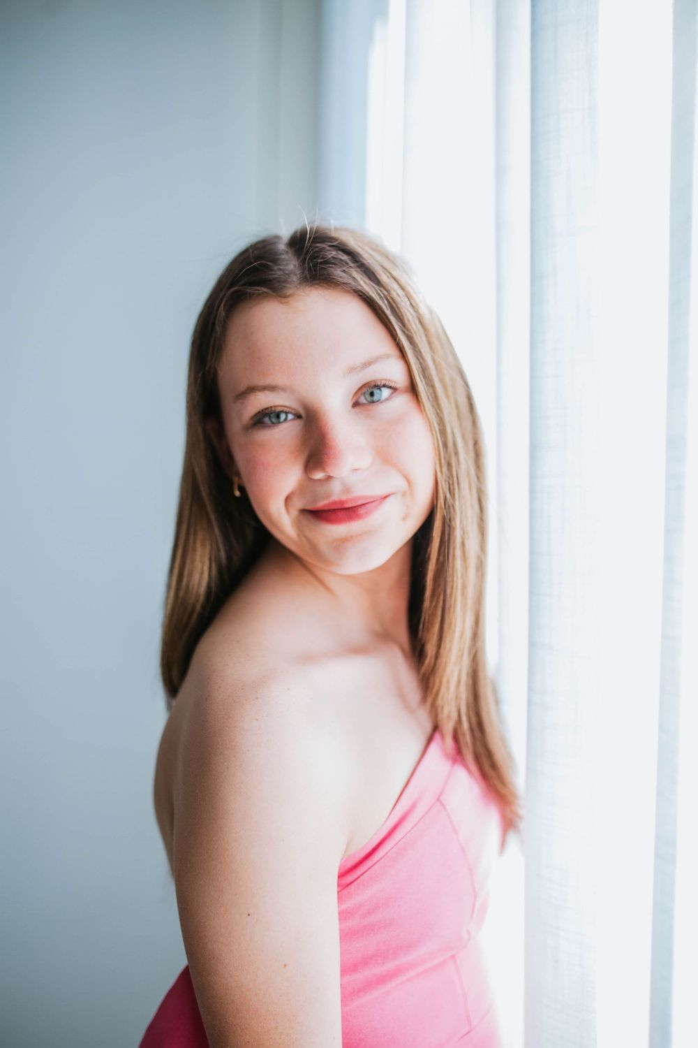 A person in a pink top poses by a window in soft natural lighting, creating a bright and airy portrait.