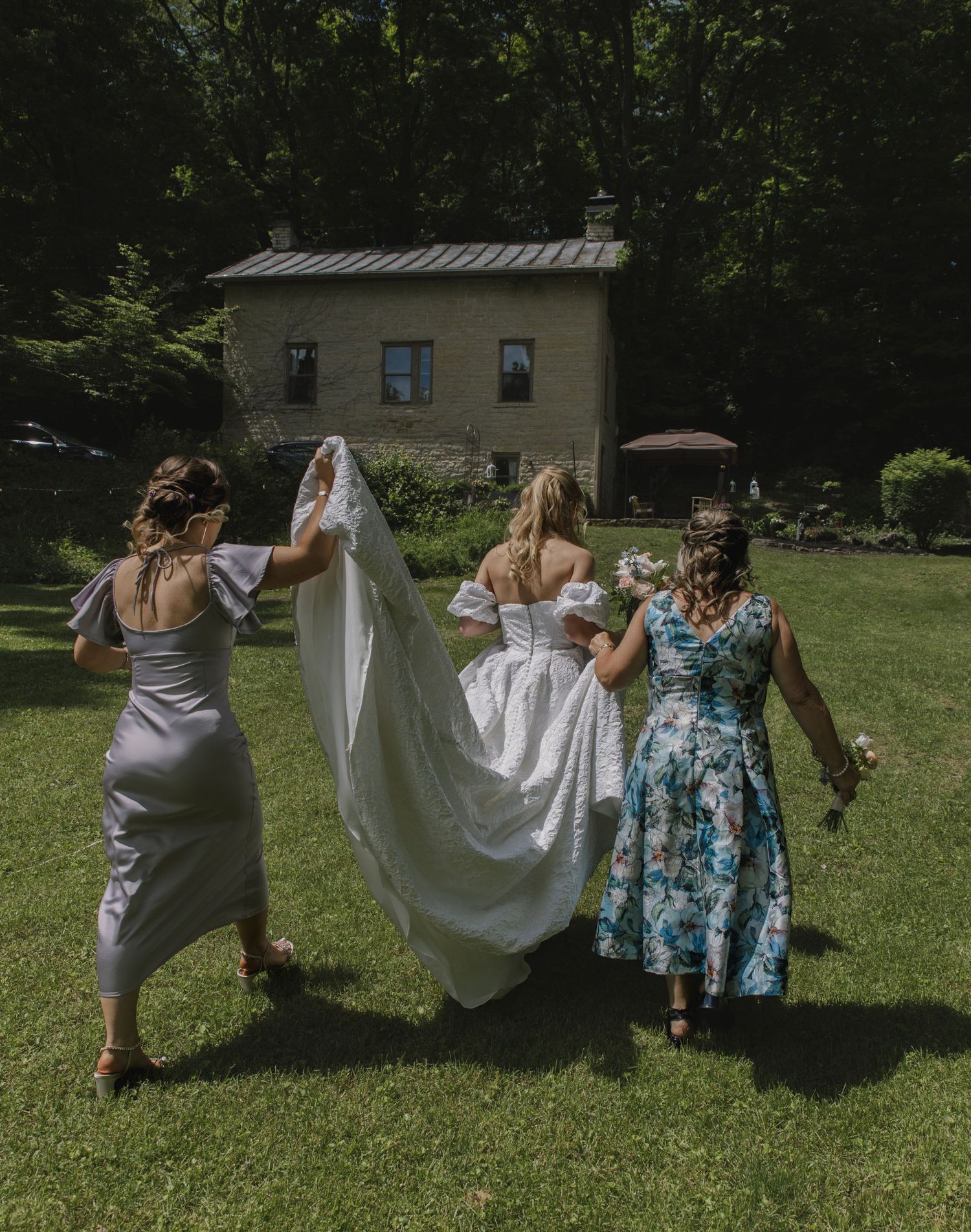Group of people lifting and arranging a white wedding dress on a sunny lawn.