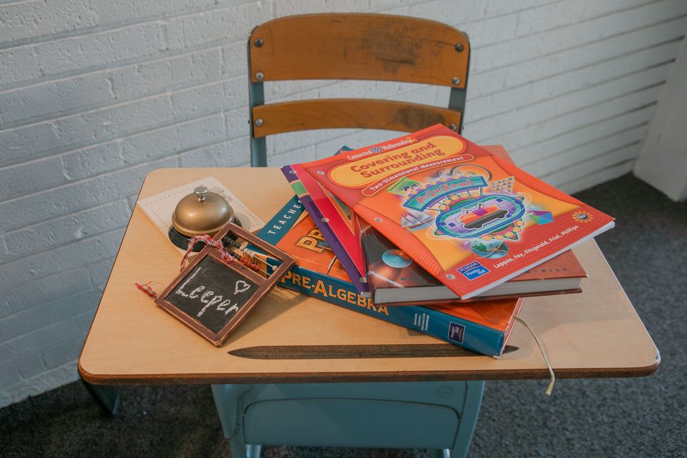 A vintage school desk with textbooks, pencils and a small chalkboard on its beige surface against white brick wall.