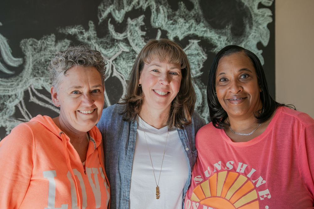 Three individuals stand close together smiling warmly against a wall with swirling black and white designs.