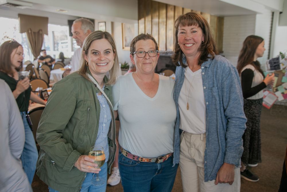 Three smiling people dressed casually stand together at an indoor social gathering with others visible in background.