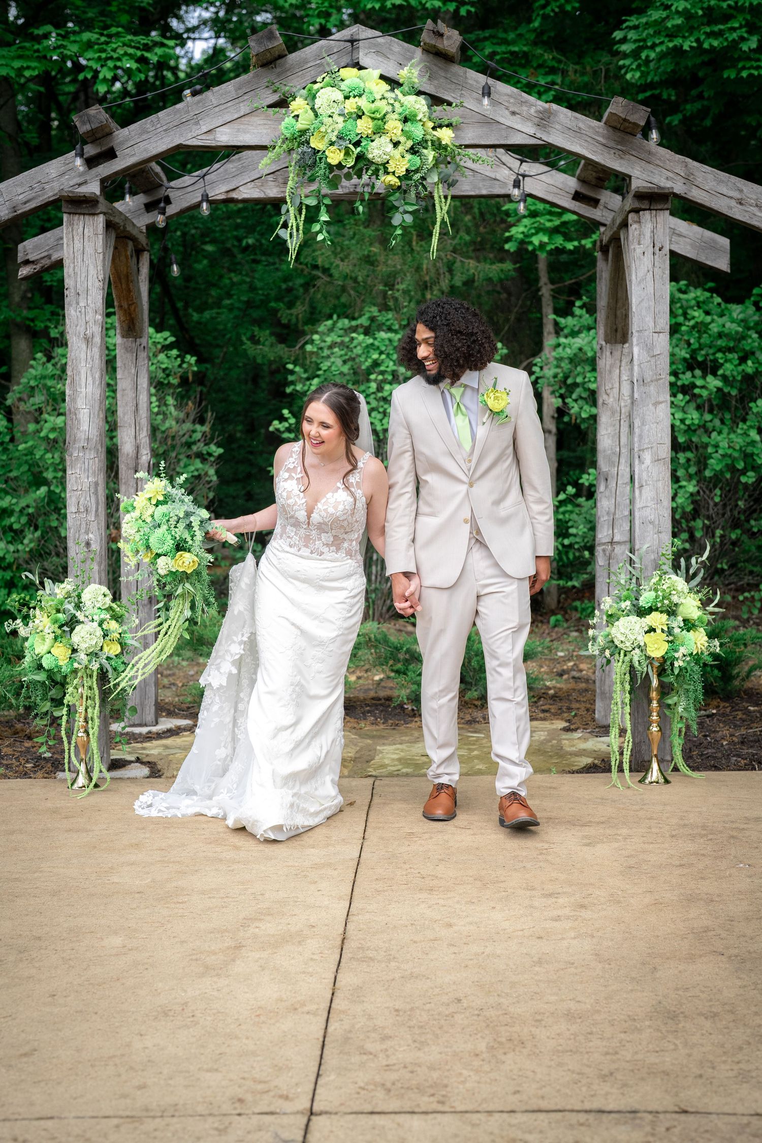 A wedding couple stands under a rustic wooden arch decorated with cascading white and yellow floral arrangements.