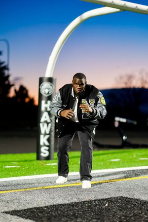 A person in a letterman jacket poses on a track field at dusk with stadium lights and Hawks signage visible.