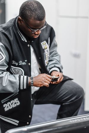 Someone wearing a black varsity jacket with 'Big Don' lettering examines a phone while sitting down.