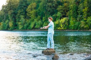 A person in light blue attire stands on a rock while fishing in a peaceful lake surrounded by lush green forest.