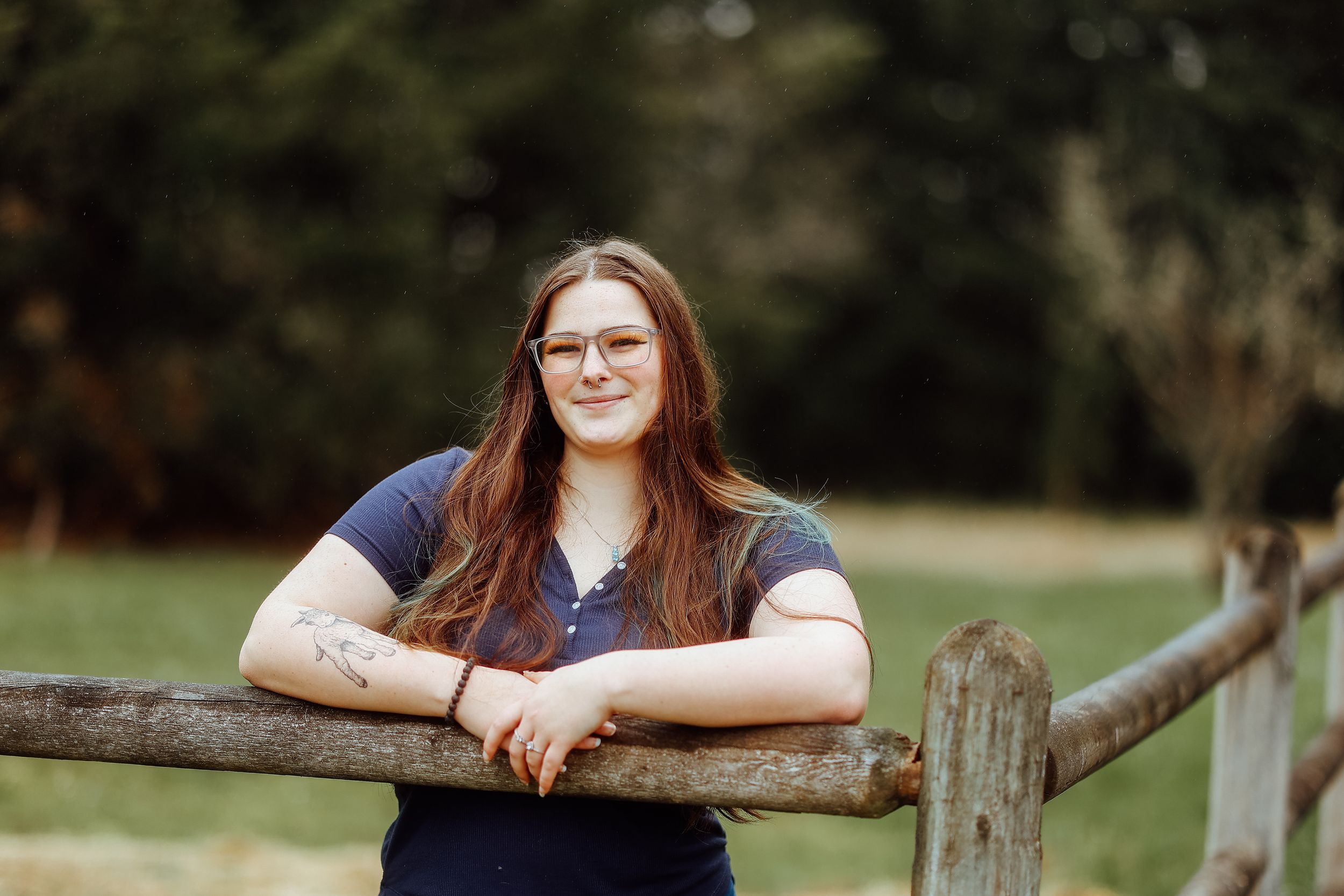 A portrait of a person with glasses in a navy shirt leaning on a wooden fence rail in a pastoral setting.
