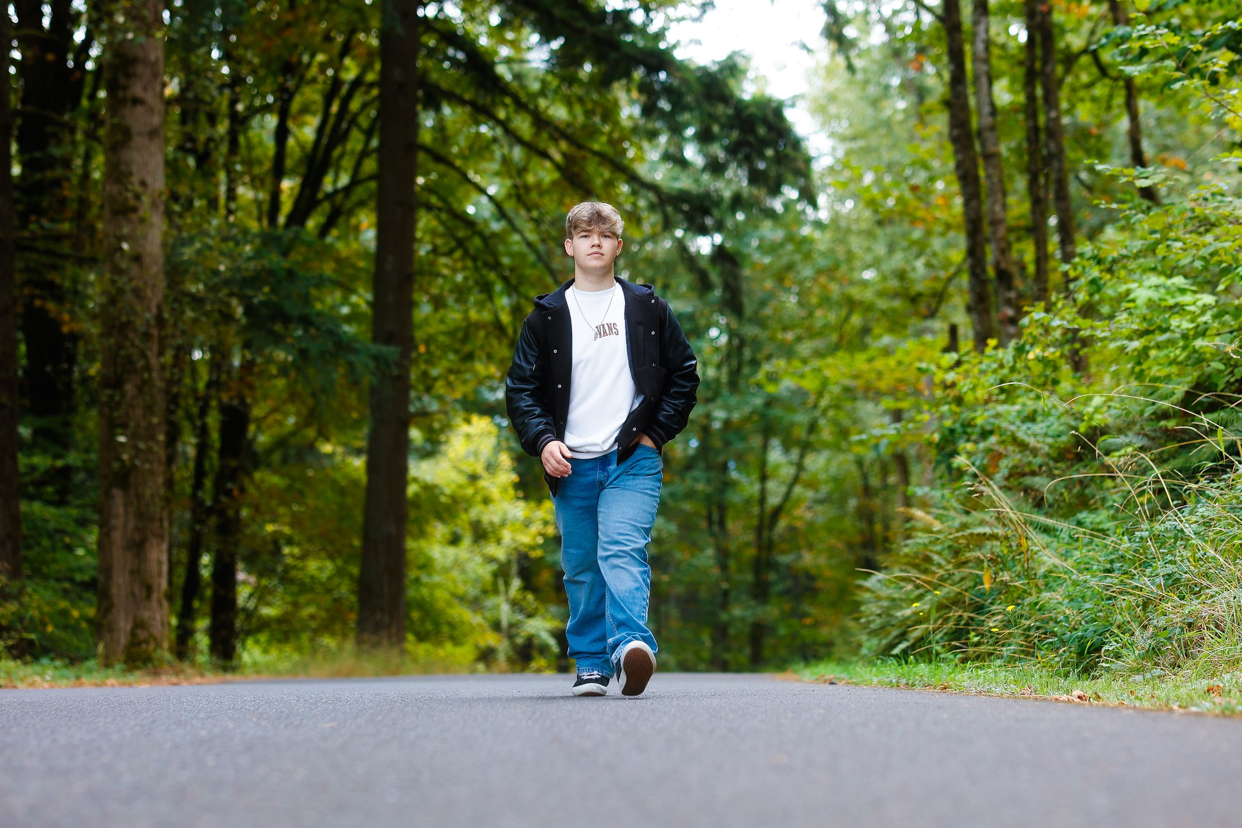 Person in blue jeans and dark jacket walking alone on a paved path through a lush green forest.