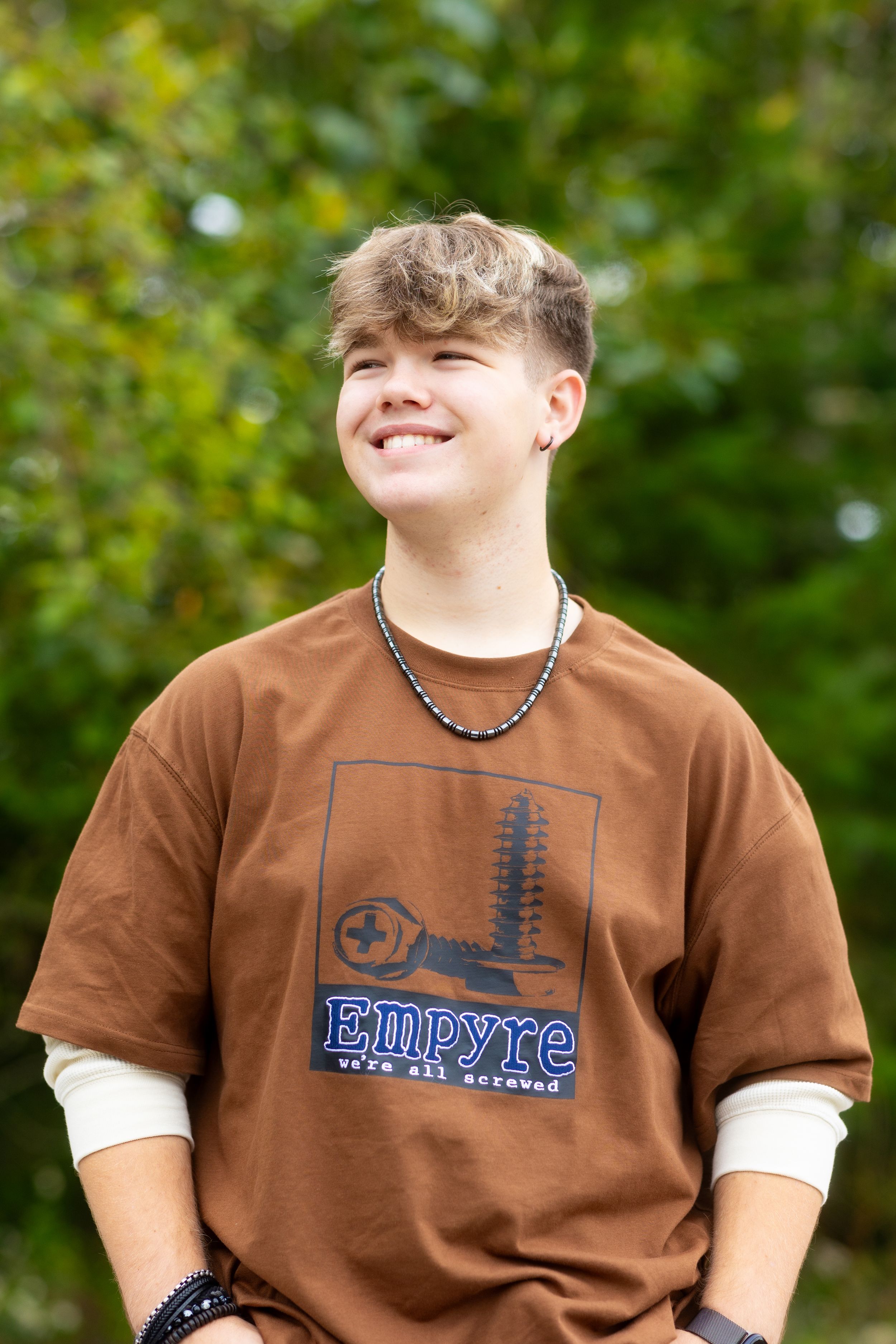 Person wearing a brown graphic t-shirt with Empyre logo and chain necklace poses outdoors against green foliage background.