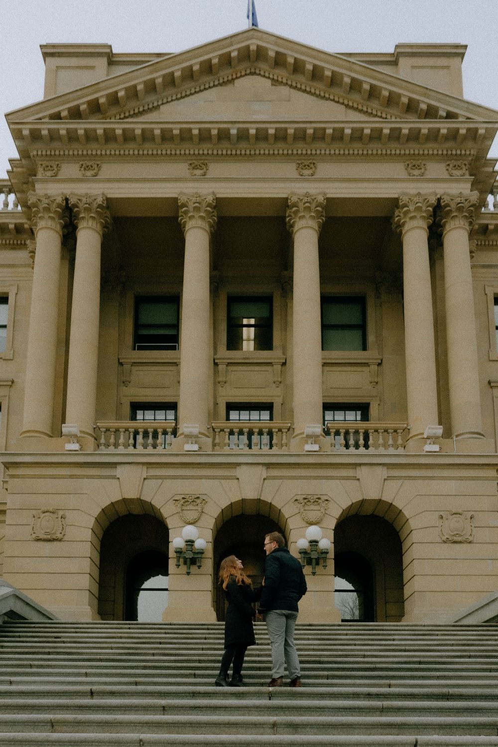 Two people stand on the steps of a neoclassical government building with grand columns and ornate architectural details.