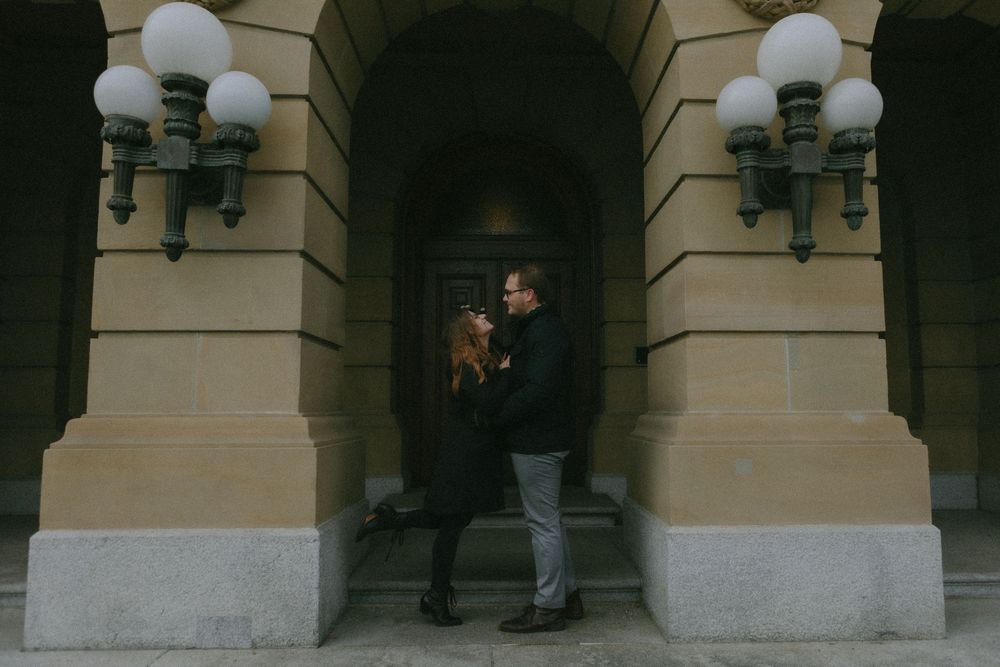 Two figures stand in a dimly lit archway entrance between ornate wall sconces at a historic stone building.