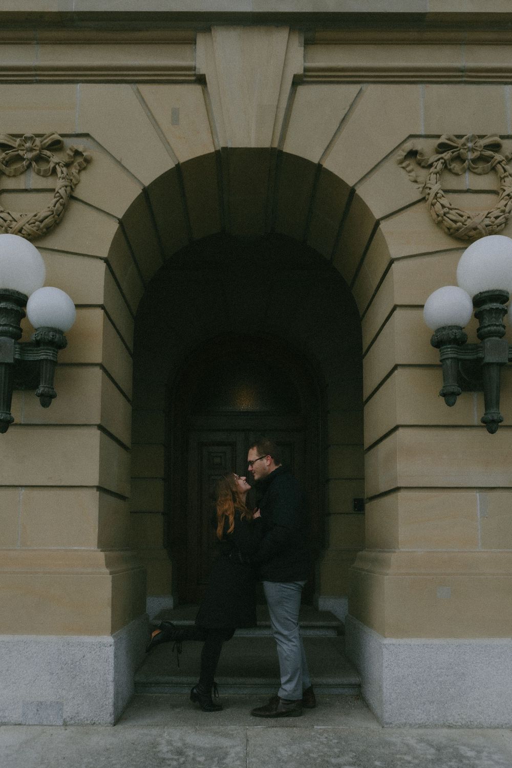 A couple shares an intimate moment in an arched stone entryway with decorative wall sconces and architectural details.