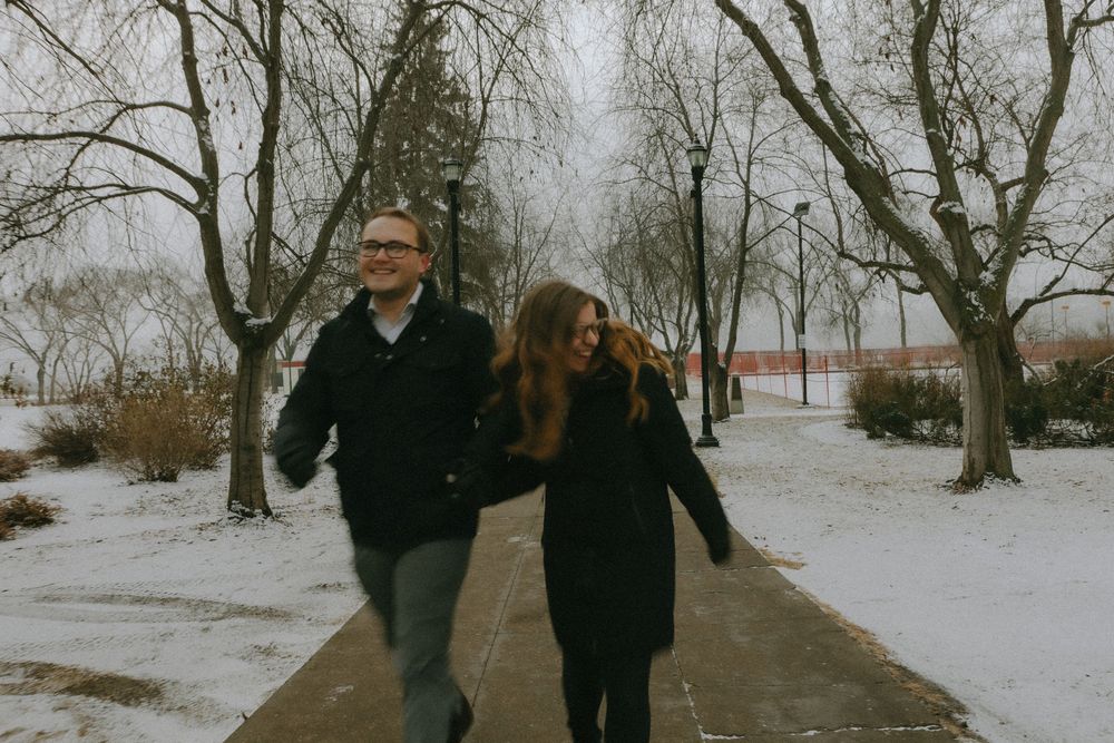 A couple walks on a snowy path through bare winter trees on an overcast winter day.