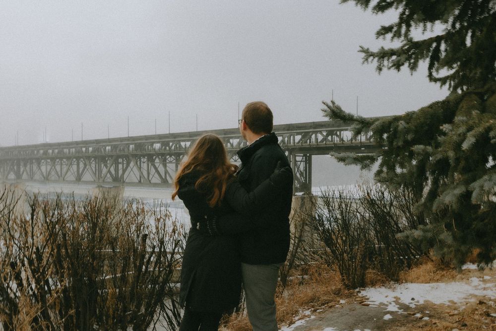 Two figures in winter coats stand overlooking a bridge on a foggy, snowy day near evergreen trees and tall grass.