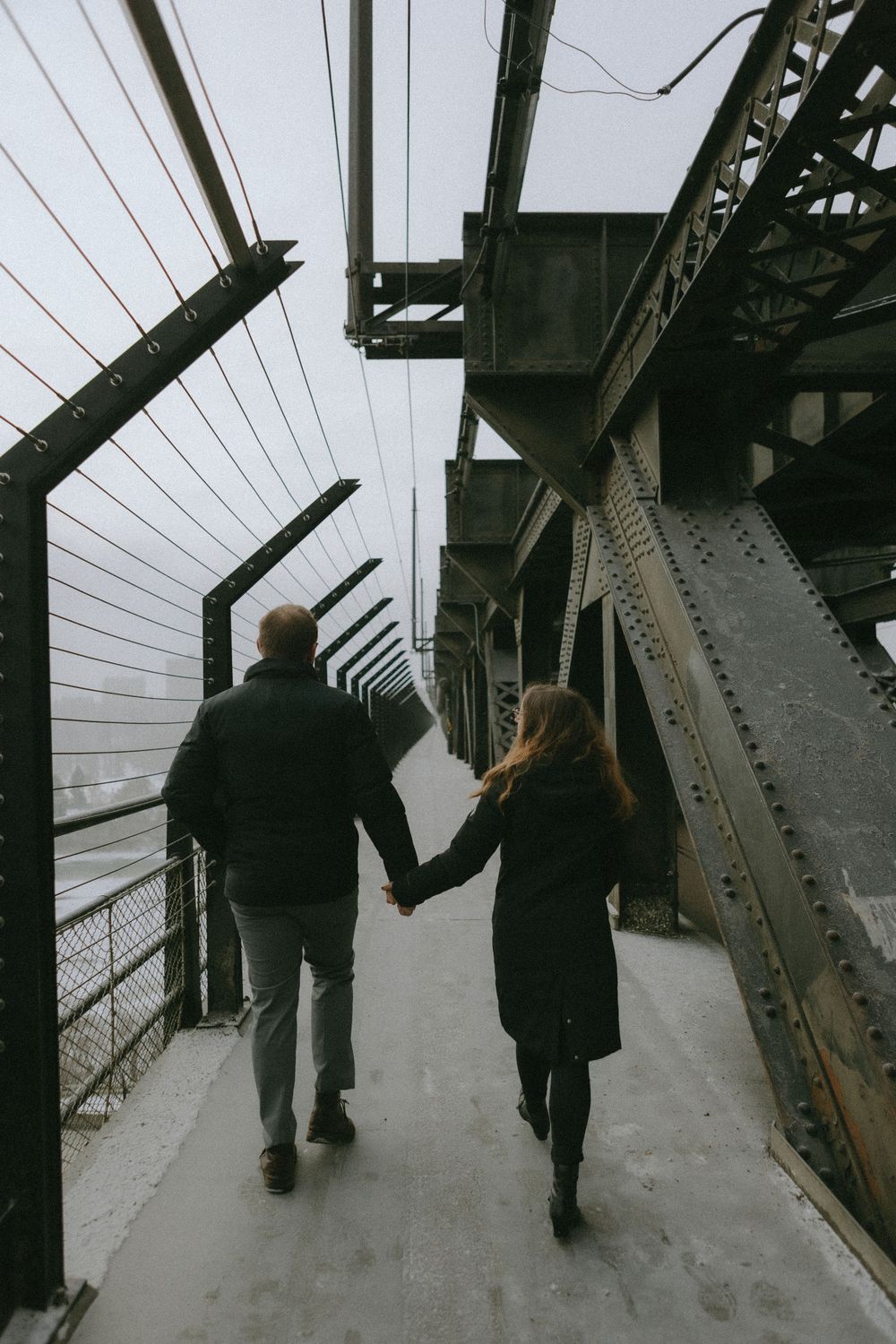 People walking along a snowy path through steel bridge trusses and beams in moody black and white photography.