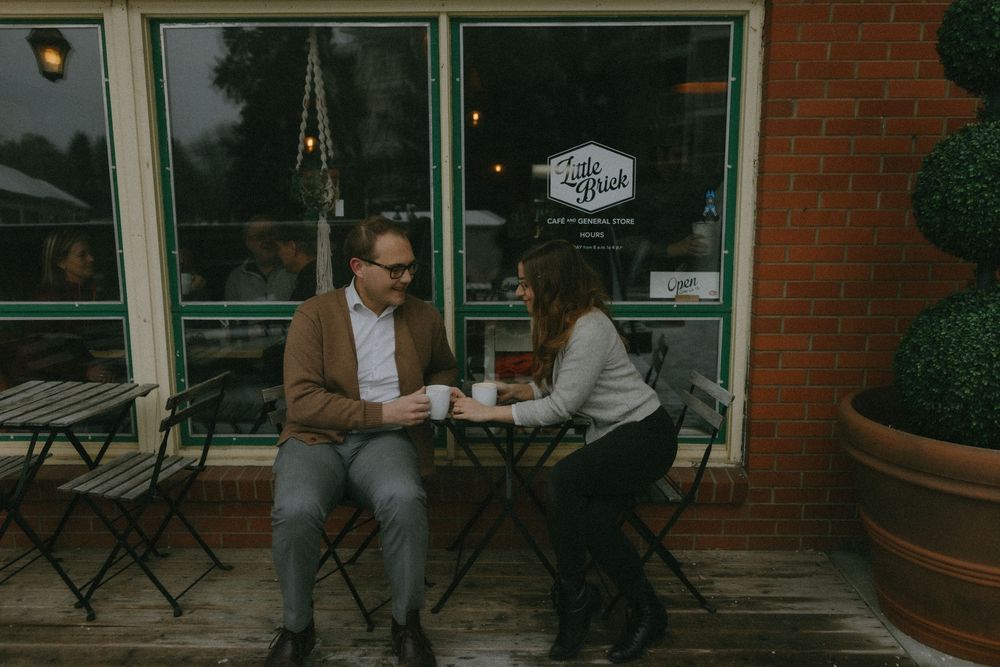 Two people sitting at an outdoor cafe table enjoying coffee and conversation in front of a brick building.