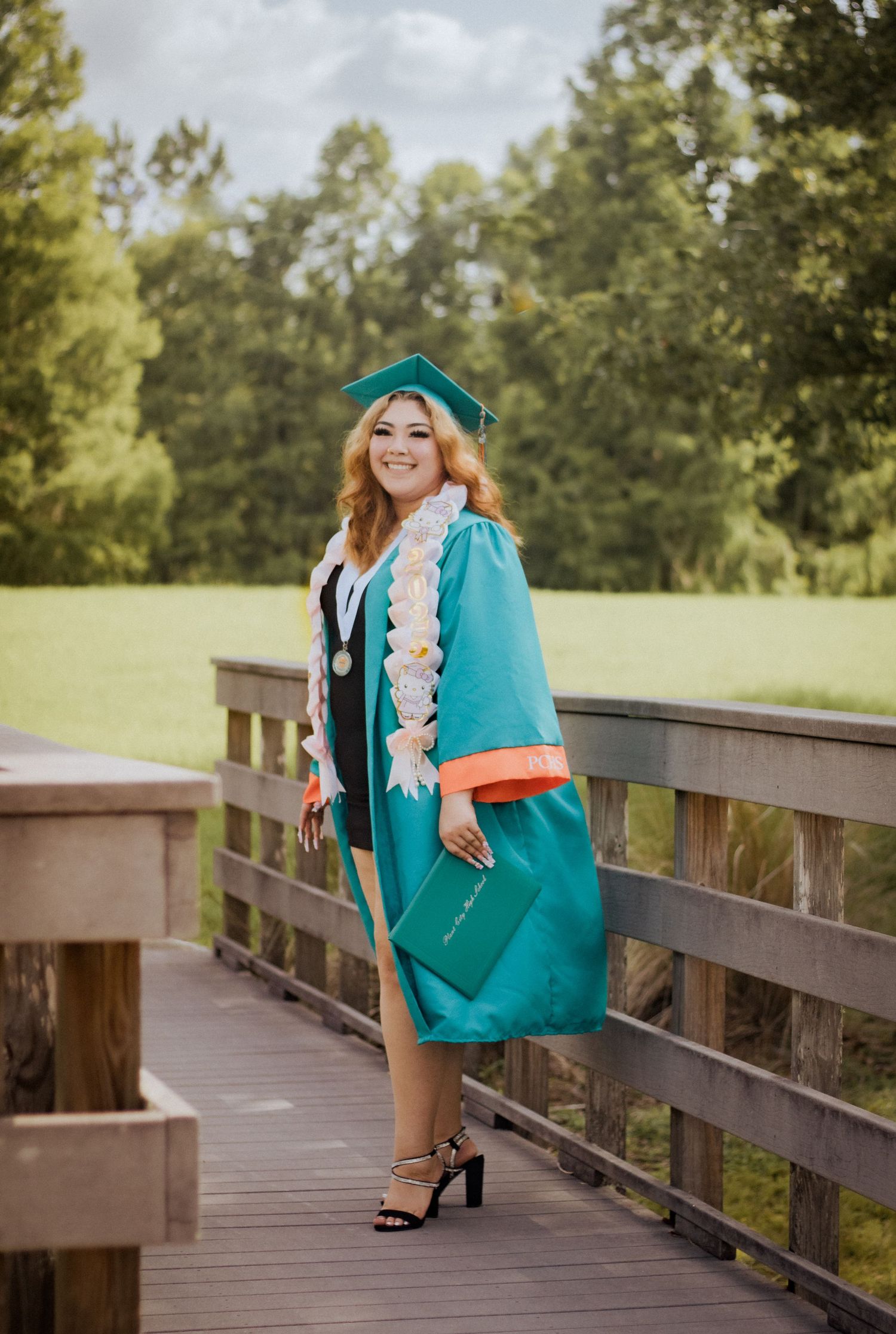 Proud graduate in teal cap and gown holding diploma on a wooden bridge outdoors.