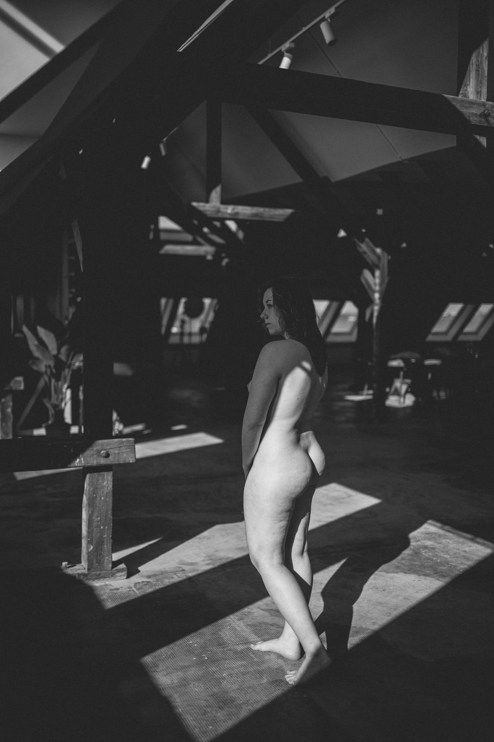 Black and white photograph of a nude figure standing in dramatic shadows beneath wooden beams in an architectural space.