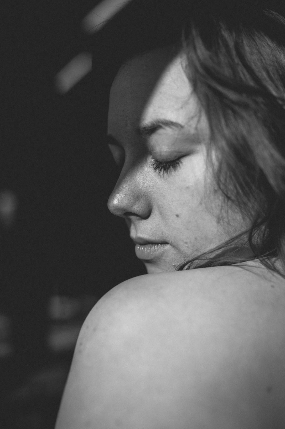 Intimate black and white portrait showing a bare shoulder and profile view with closed eyes against dark background.