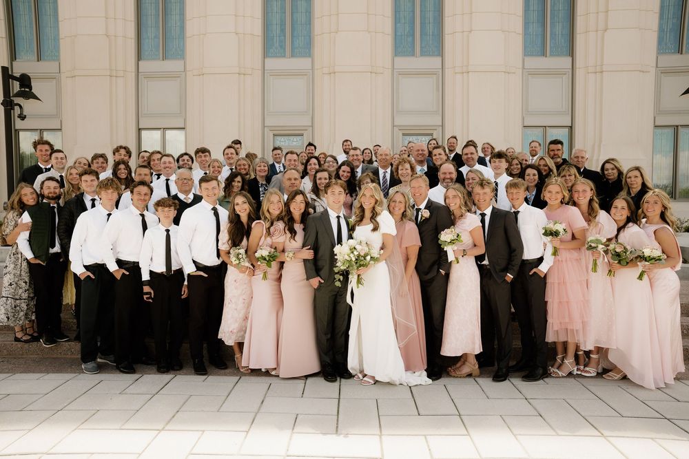 Wedding party members gather for formal group photo wearing pink bridesmaids dresses and black tuxedos.
