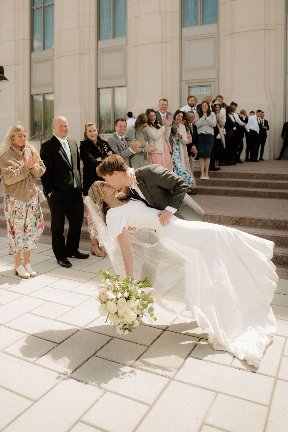 A romantic wedding dip kiss moment with guests watching from the church steps.