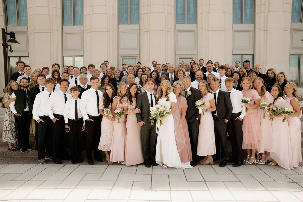 Large wedding party poses together in formal attire, with bridesmaids in pink dresses and groomsmen in black and white.
