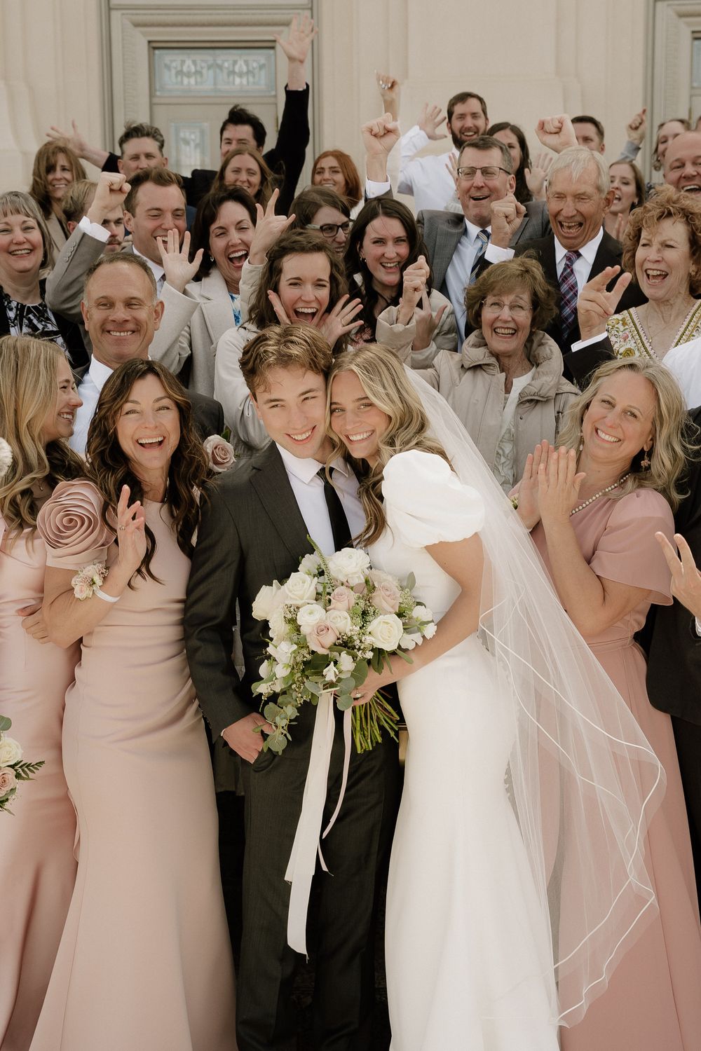 A joyful wedding party poses together with the newlyweds at the center, bridesmaids in blush pink dresses surround them.