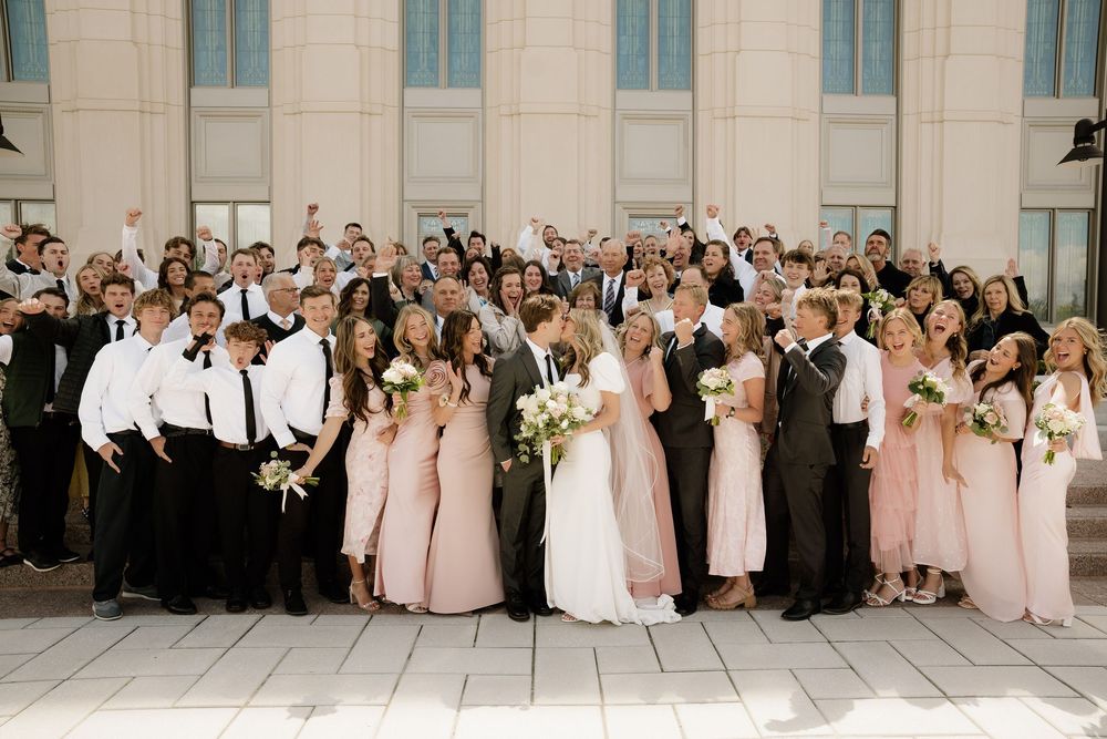 Large wedding party in black and pink attire poses together on church steps for group photo.
