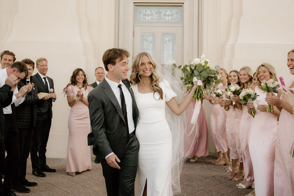 Newlyweds walk past wedding party members in pink dresses and dark suits.
