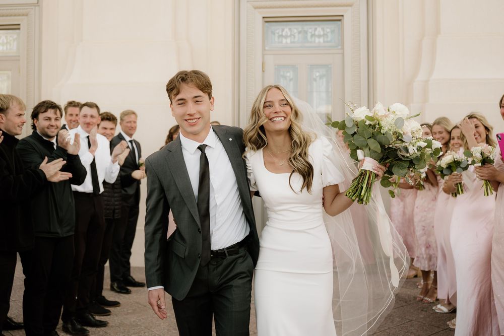 Newlyweds exit church ceremony between rows of guests, bride in white dress holding floral bouquet while groom wears black suit.