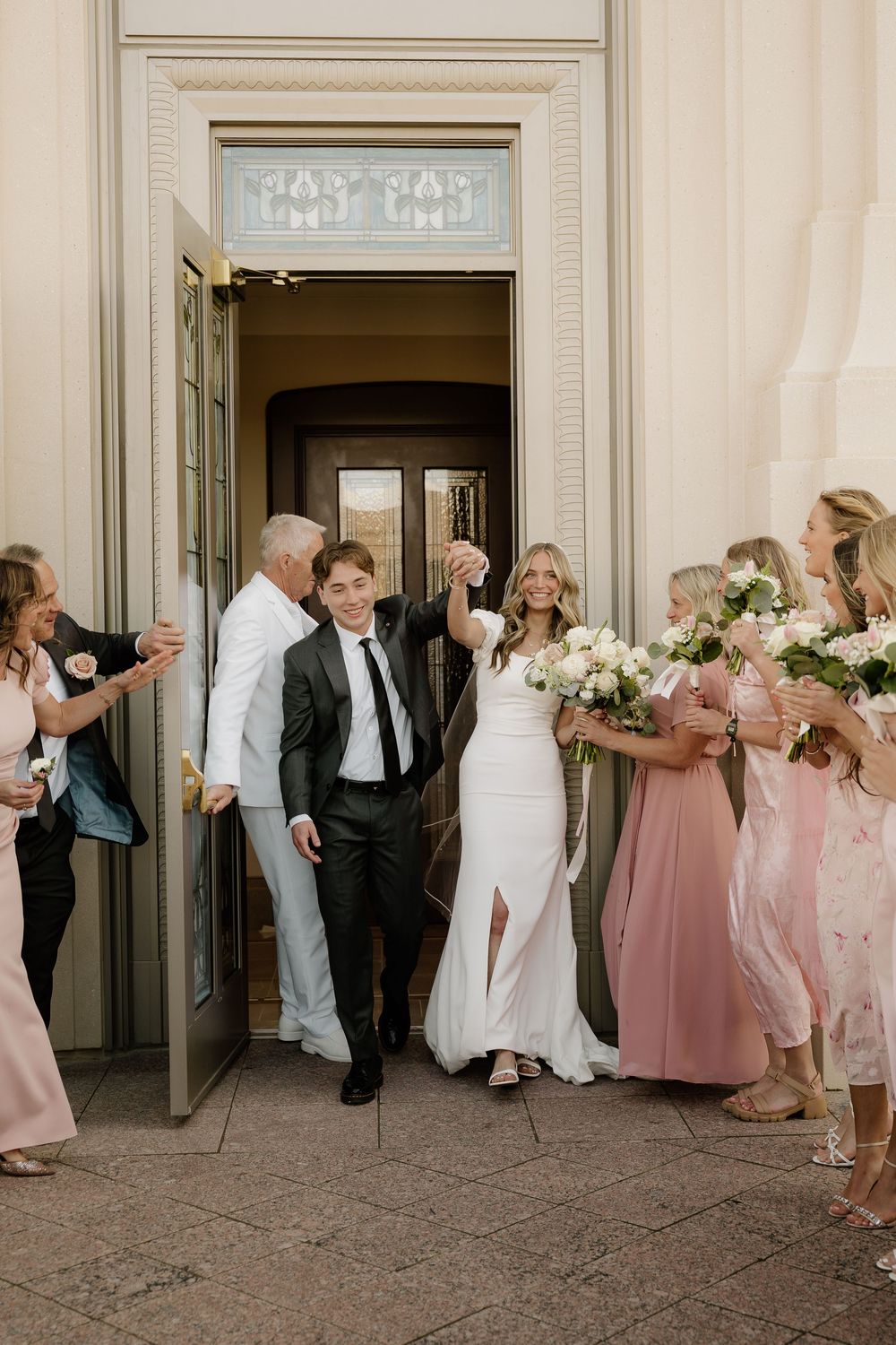 Wedding party in black suits and blush pink dresses lean playfully against a classic cream-colored building exterior.