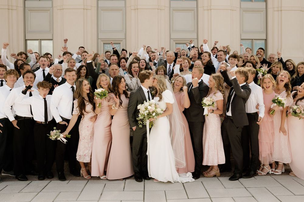 Large wedding party in formal attire including pink bridesmaid dresses and classic black suits poses together on light stone patio.