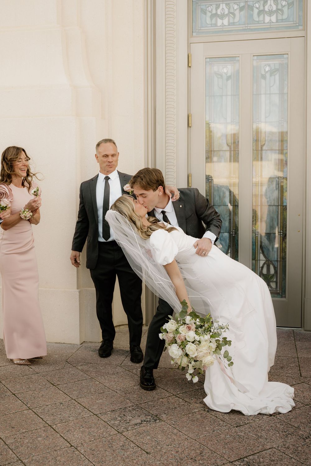 A romantic wedding moment outside an ornate glass doorway with pink and white floral bouquet details.