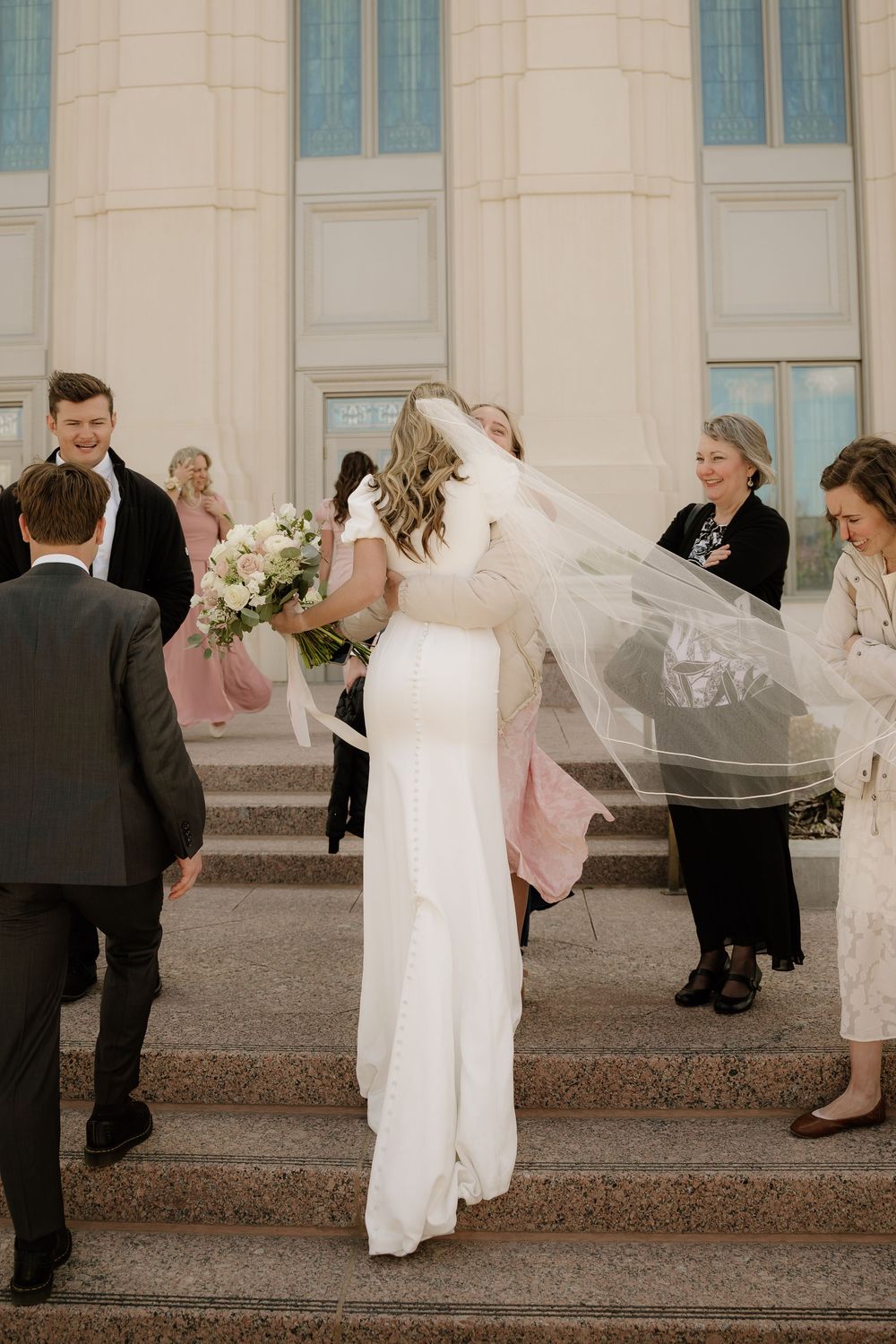 A bride in a flowing white dress and veil stands on church steps surrounded by wedding guests after the ceremony.
