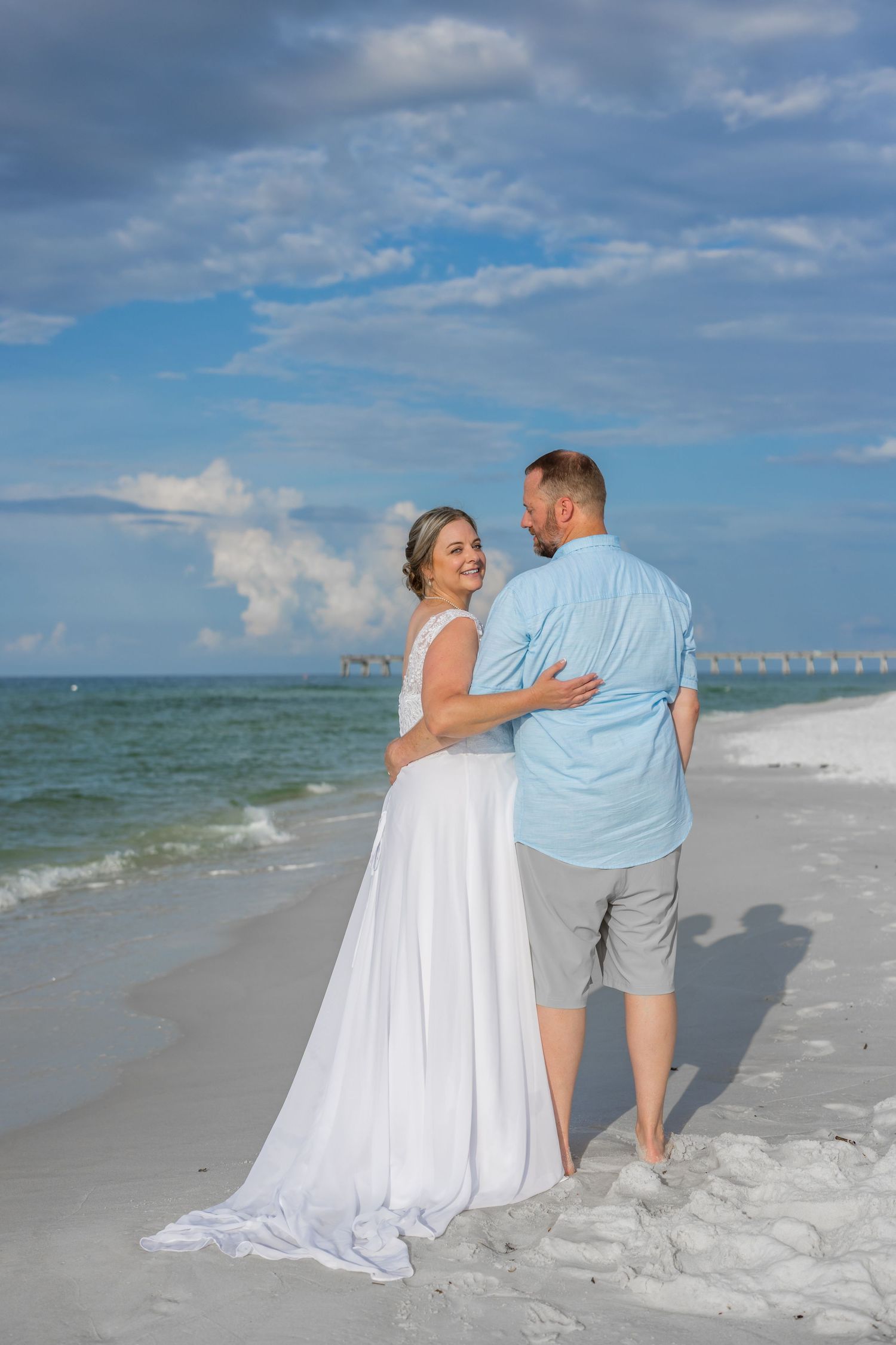 A couple in white and light blue clothing walking together along a pristine sandy beach with ocean waves and blue sky.