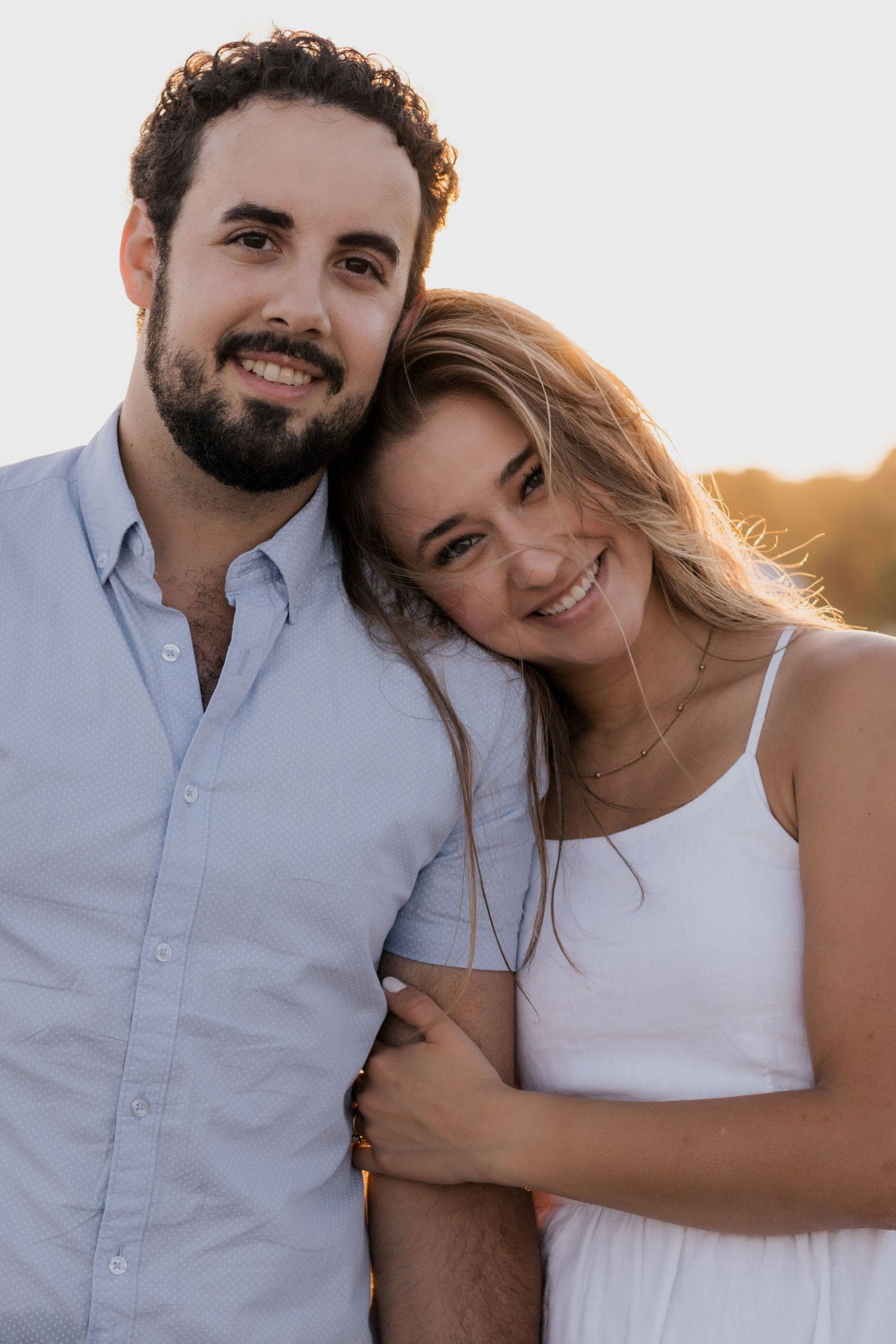 Smiling couple embraces during golden hour sunset, woman in white dress leans on mans shoulder.