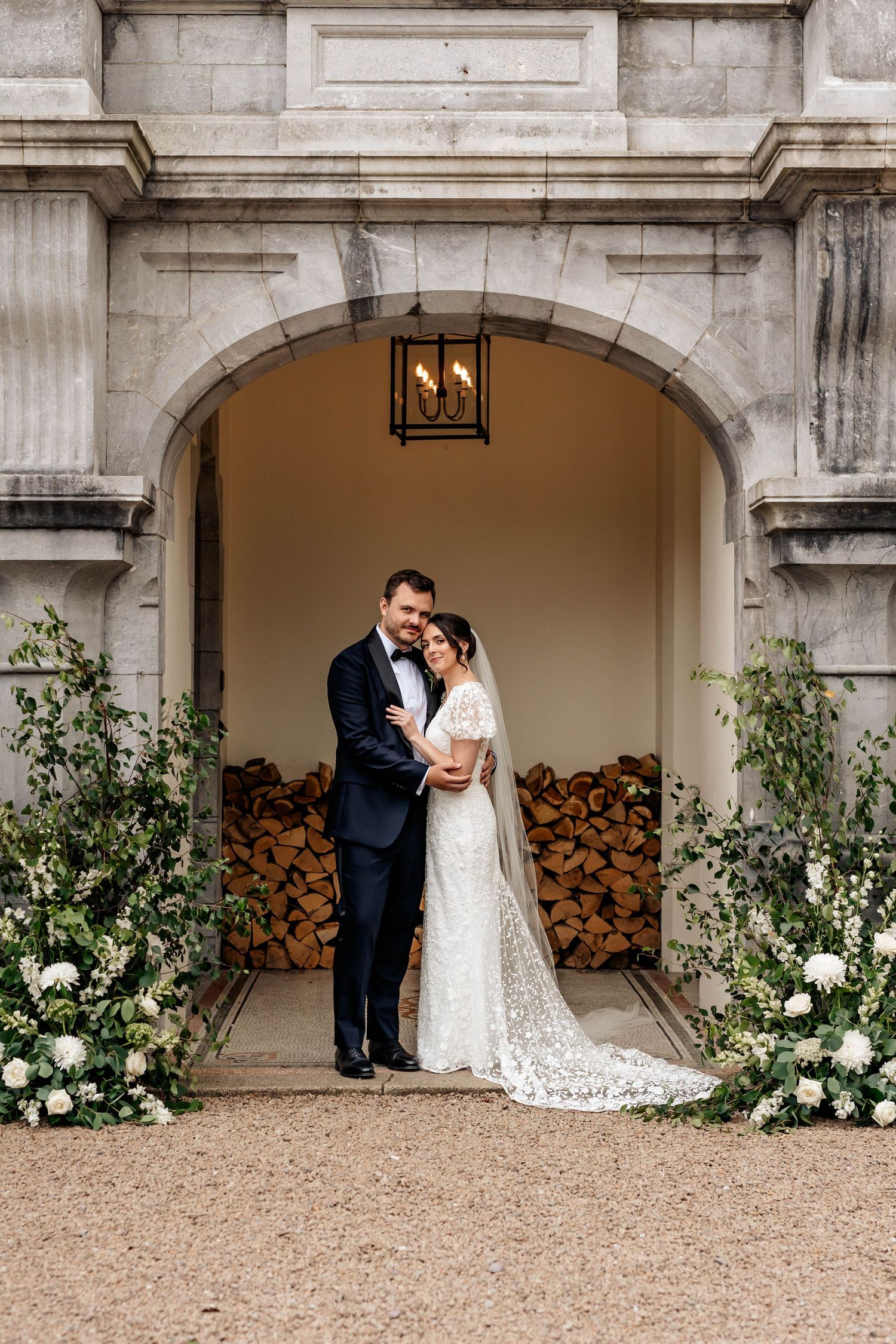 A wedding couple poses under a stone archway decorated with white floral arrangements.