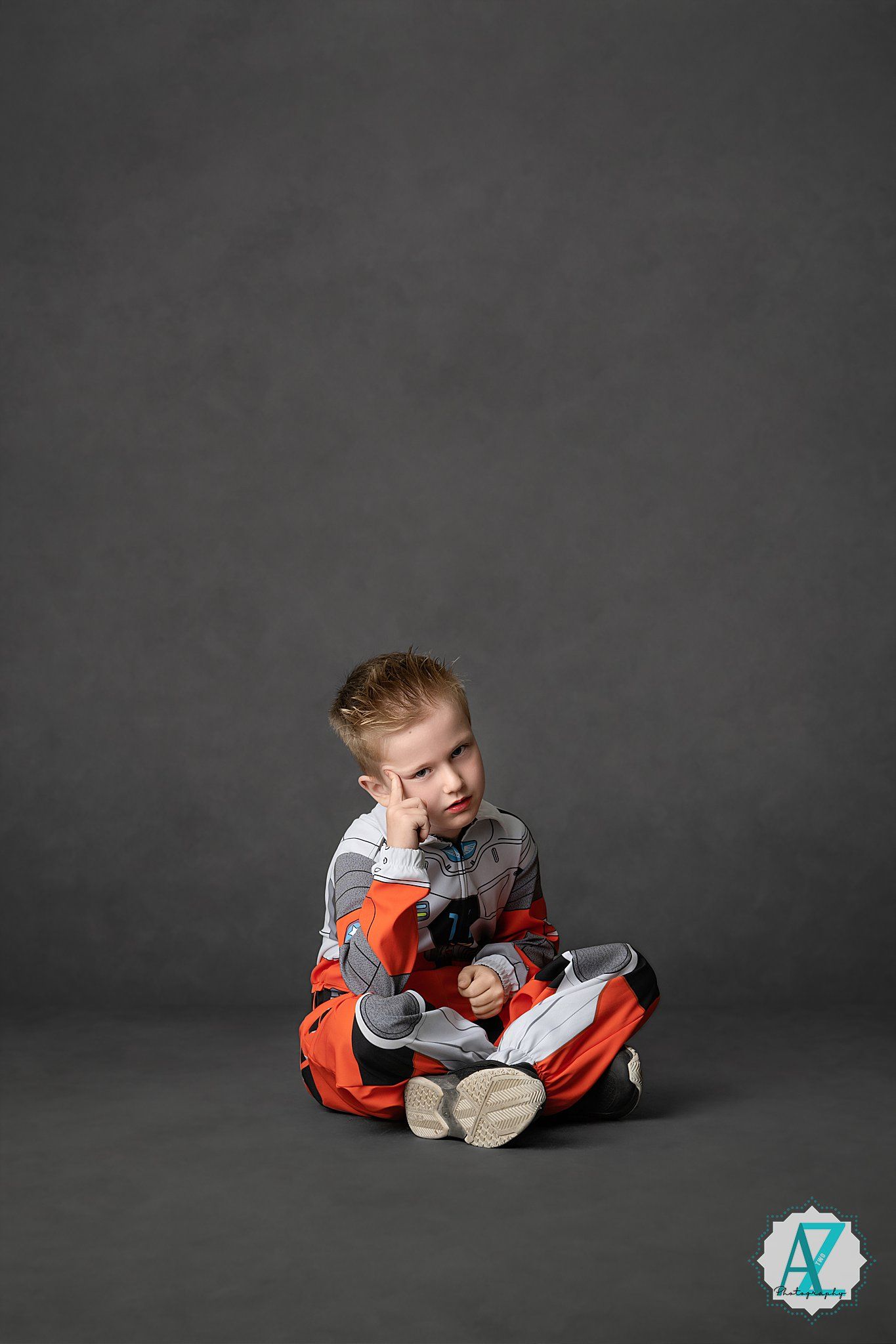 Small child in orange and gray outfit sitting cross-legged on dark floor looking pensive against gray background.