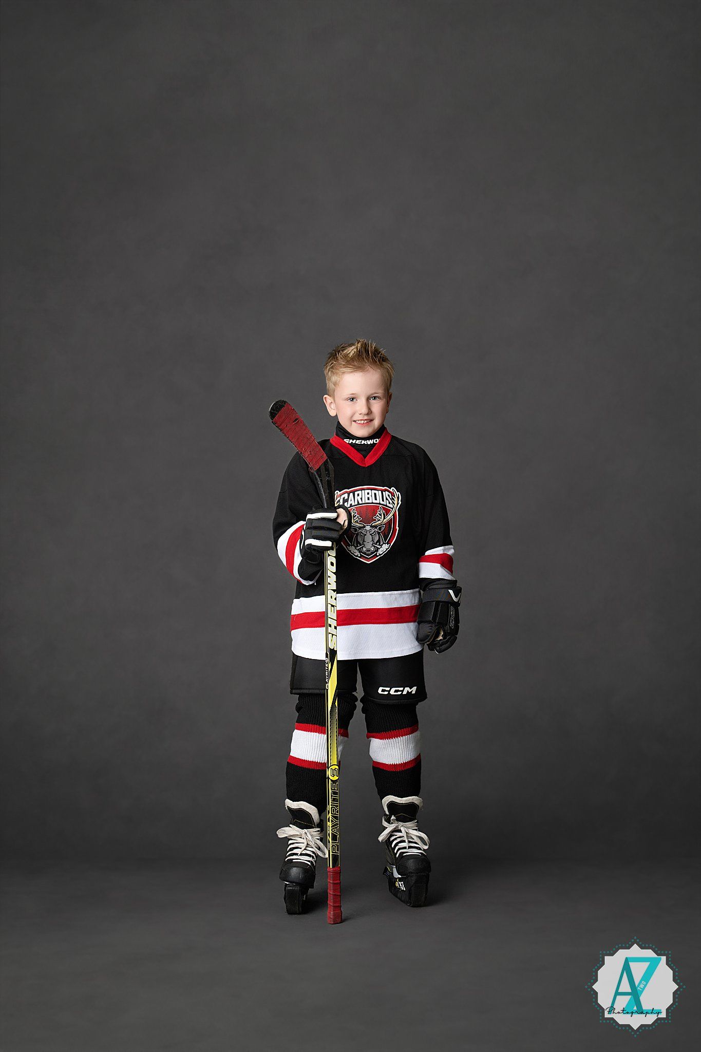 Young hockey player in black and red uniform stands on skates holding a red hockey stick against a dark background.