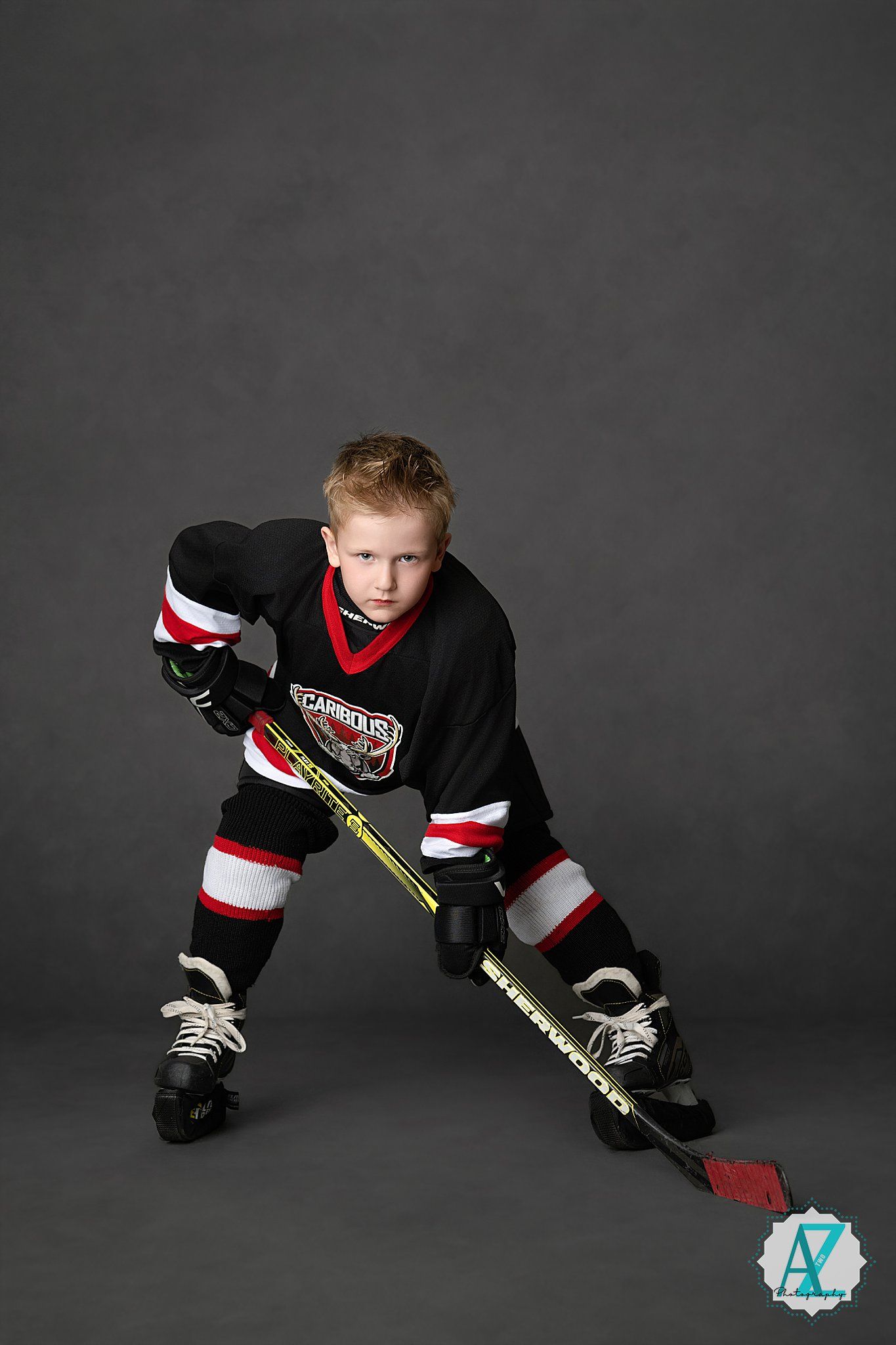 Young hockey player in black jersey and protective gear poses in ready stance with hockey stick on dark background.