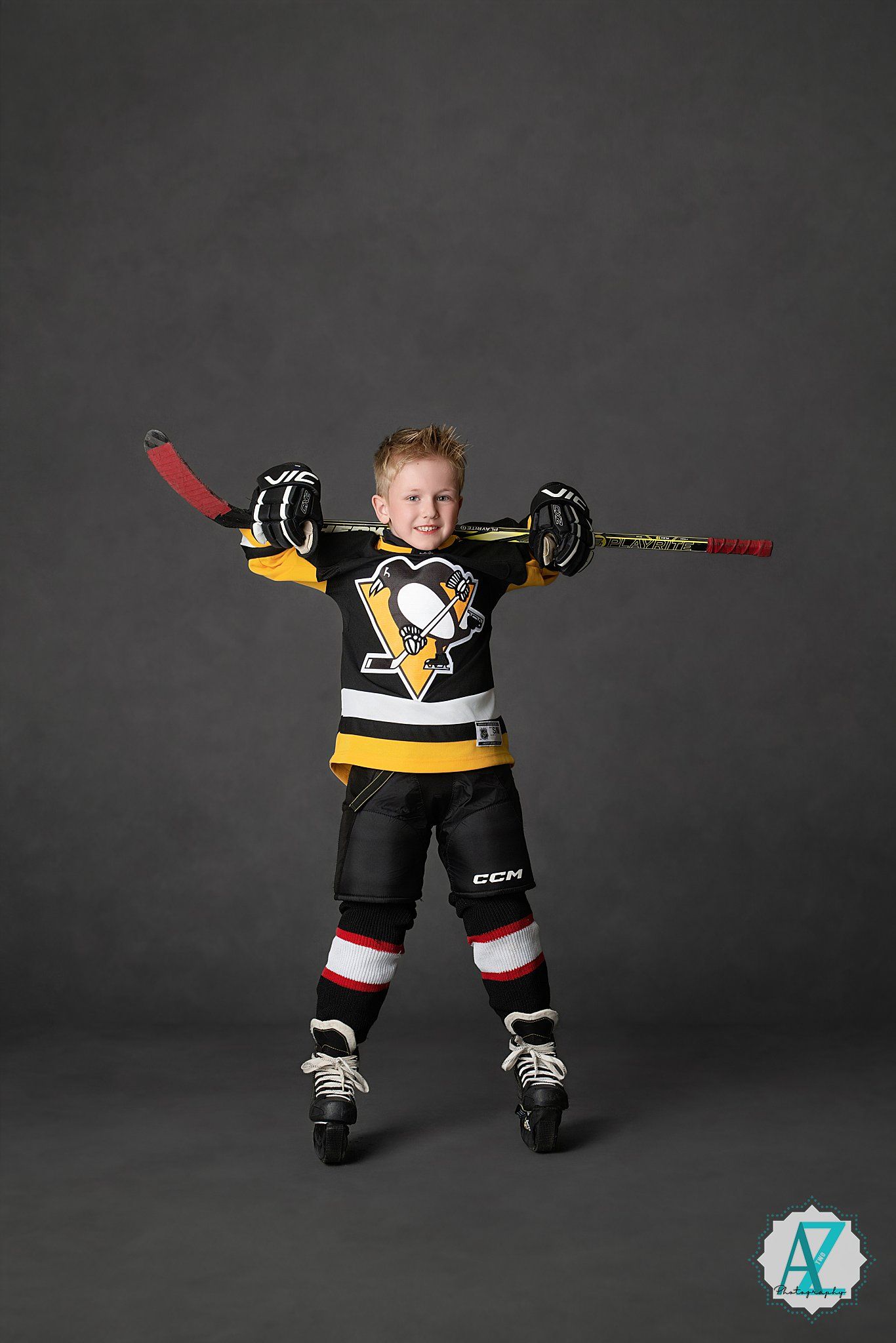 Young athlete poses in Pittsburgh Penguins hockey uniform with stick raised against dark background.