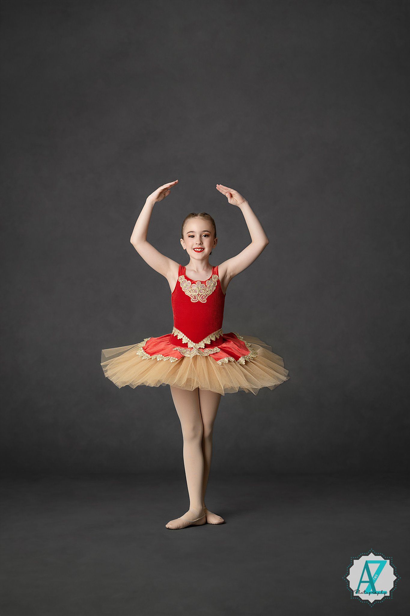 Young ballerina poses gracefully in a red and gold classical tutu against a dark studio backdrop.