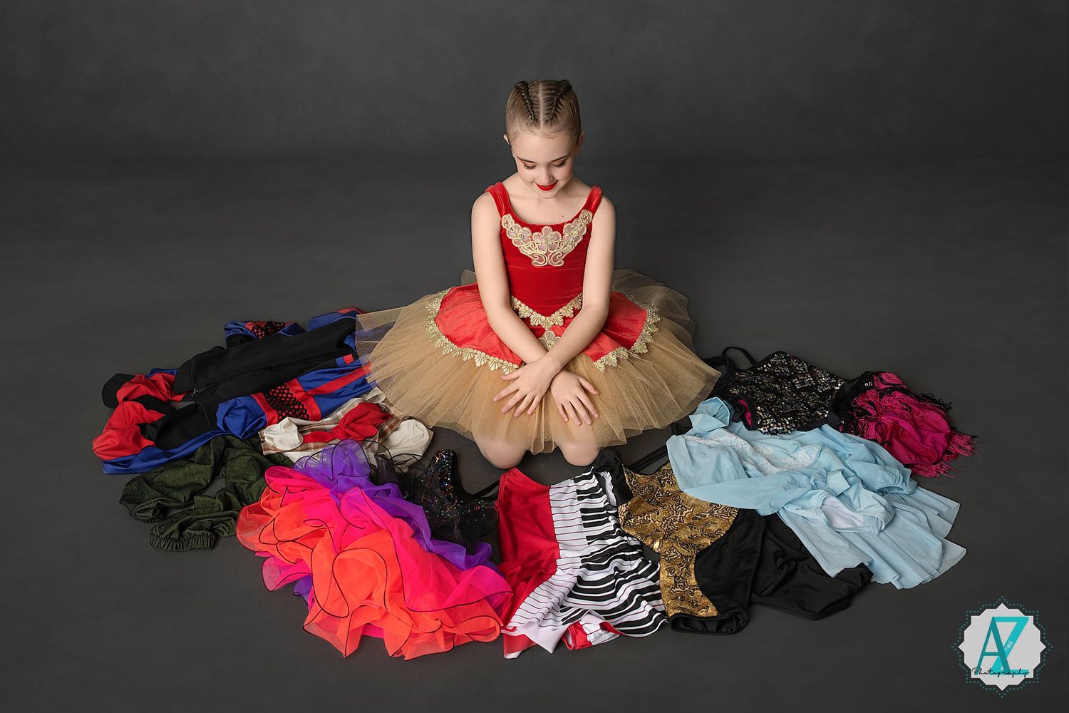Ballet dancer wearing red and tan tulle dress poses with various dance outfits arranged in a circle.