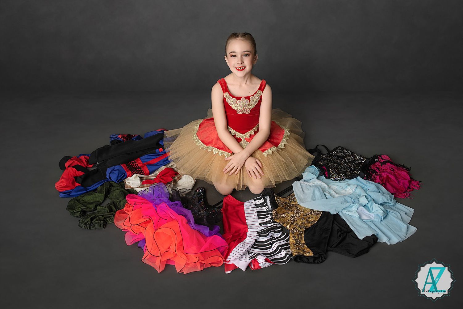 Dancer in red and beige tutu sits surrounded by colorful dance costumes on dark backdrop.