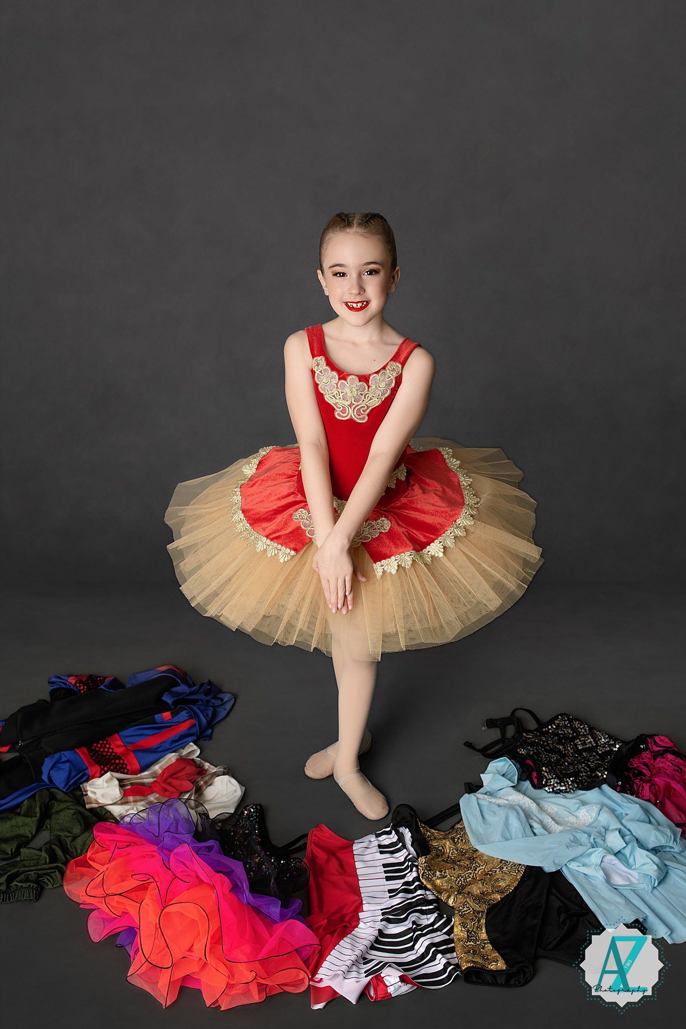 Professional ballet dancer in red tutu poses gracefully among colorful dance costumes scattered on dark floor.
