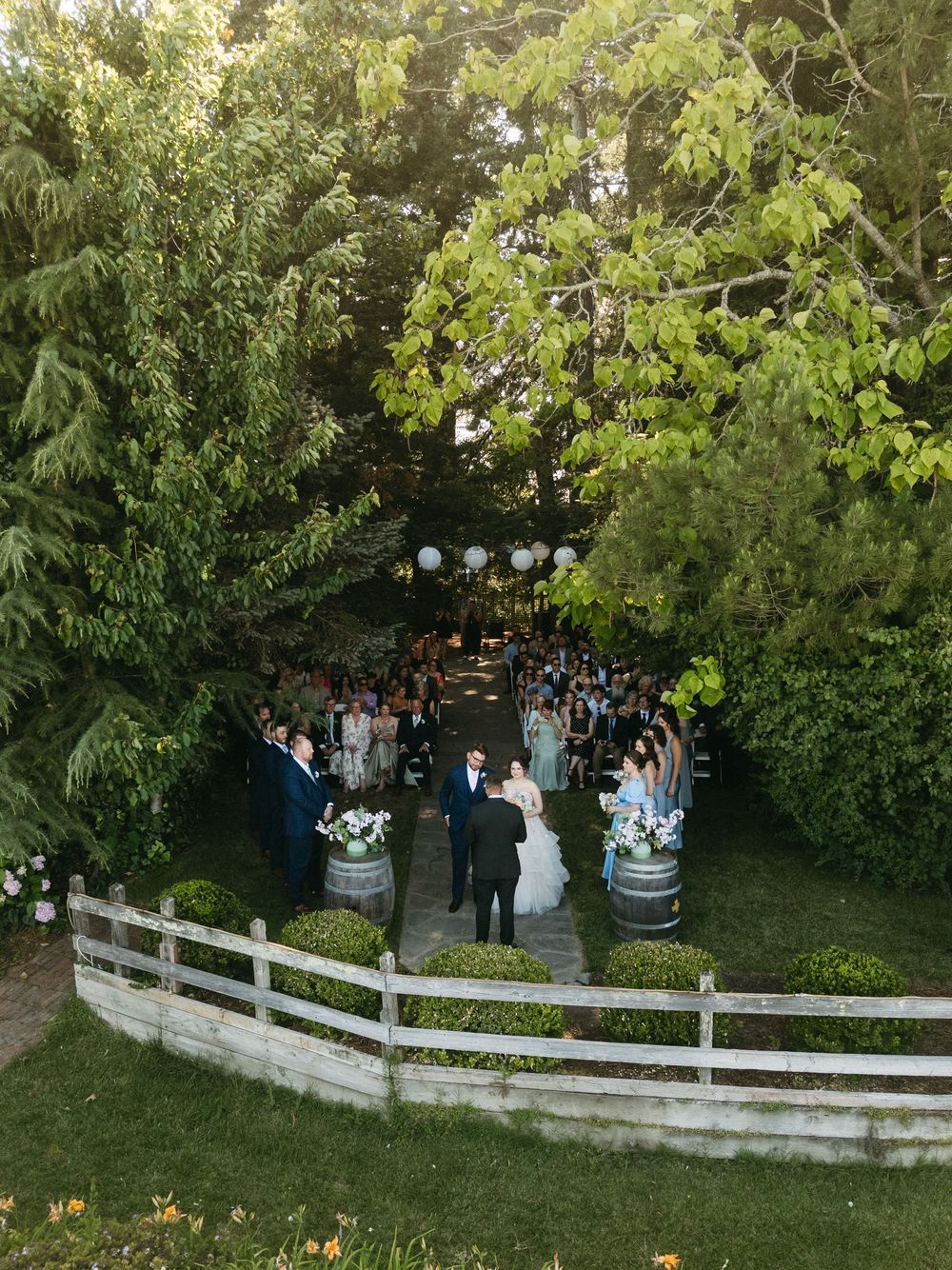 Aerial view of an outdoor wedding ceremony surrounded by lush green trees, with guests gathered along a white fence.