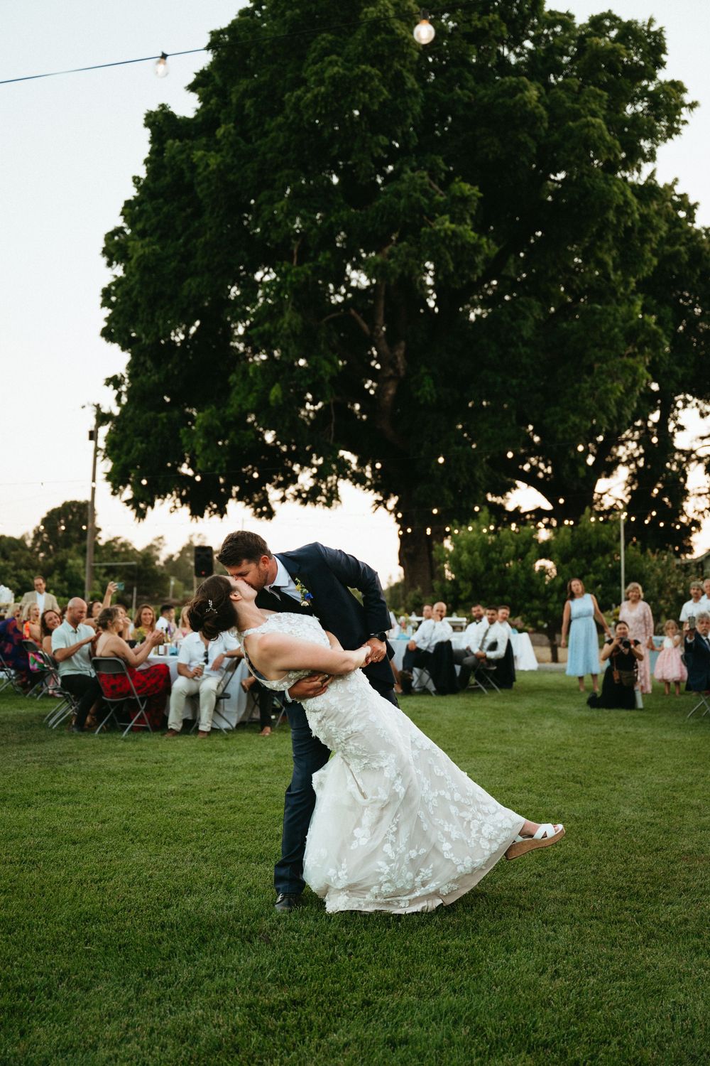 A romantic first dance unfolds on a lawn at an outdoor garden wedding reception under a large shade tree at sunset.