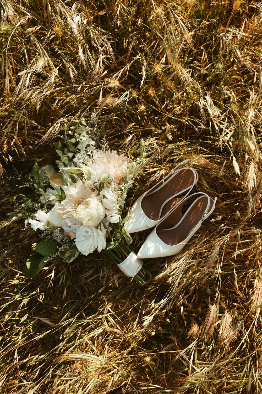 A pair of elegant white bridal shoes with floral decorations arranged on dried grass or straw.